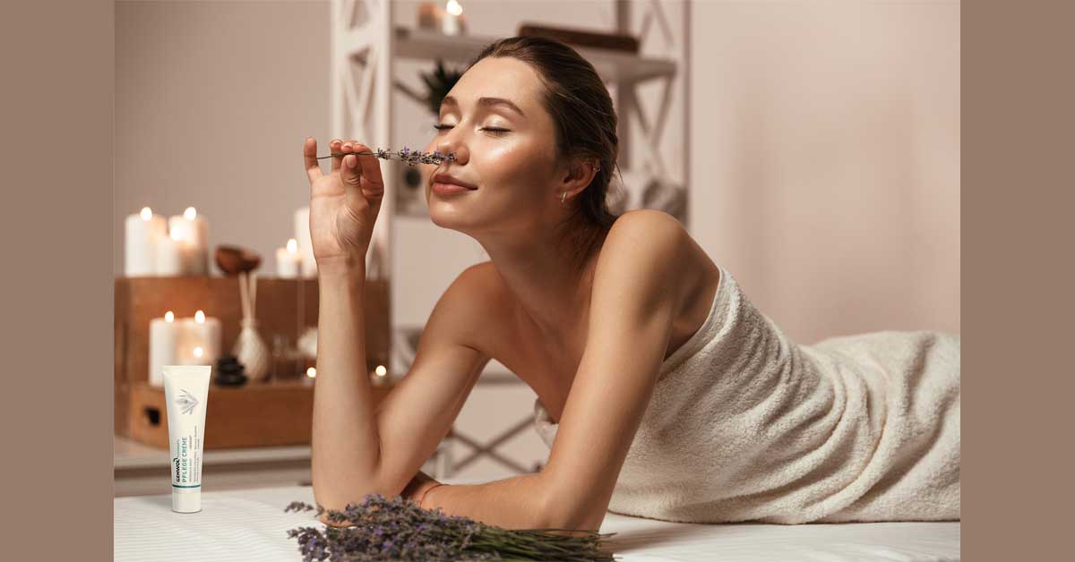 A woman wrapped in a towel is smelling lavender and has a Gehwohl cream tube placed beside her. 