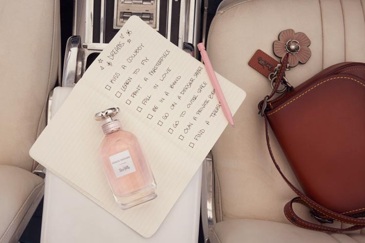 The behind the scenes image from the making of the campaign shows the 'Coach Dreams' flacon - referencing vintage apothecary bottles, with to-do list starting with "Kiss A Cowboy" for the visualization of a sunny trip through the American West. Beside the perfume bottle at a car's front seat, a signature leather saddle bag by Coach. 