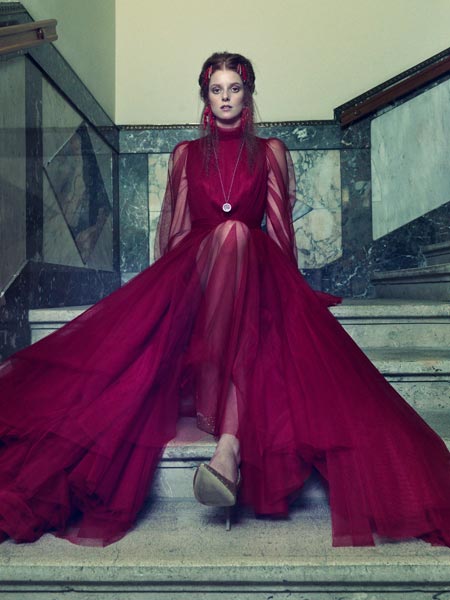 A model sits at a staircase dressed in a red semi-transparent evening gown by Thang de Hoo Couture